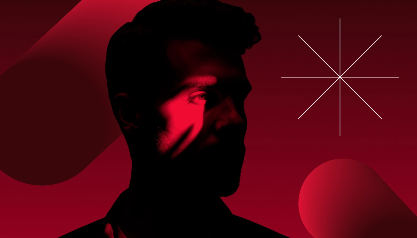 A cybercriminal stands in the shadows against a red background, his eye spotlit